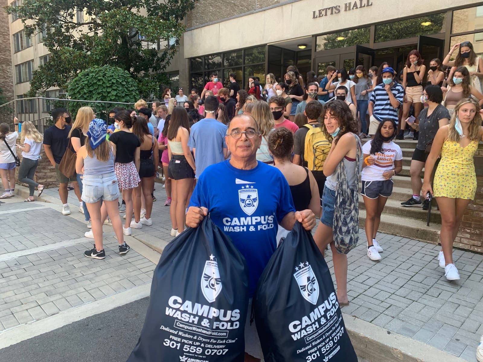 Campus Wash and Fold staff member holding two branded laundry bags outside Letts Hall at American University in Tenleytown, Washington DC