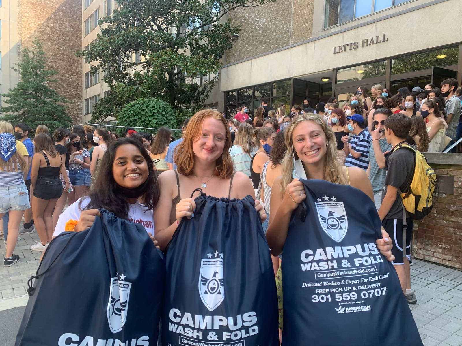 American University students smiling and holding Campus Wash and Fold branded laundry bags outside Letts Hall in Tenleytown, Washington DC